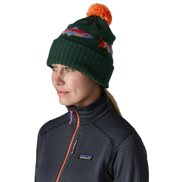 Person wearing a green Powder Town Knit beanie with a colorful pom-pom and salmon designs, and a dark jacket with a Patagonia logo over a white background.