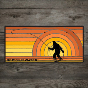 A Rep Your Water Sunset Squatch Sticker, featuring a bigfoot fishing silhouette, on a wood grain background.
