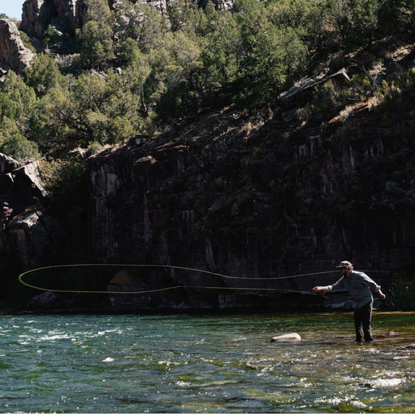 Person fly fishing with the Sage Arrow Fly Rod in a river with rocky cliffs and trees in the background.