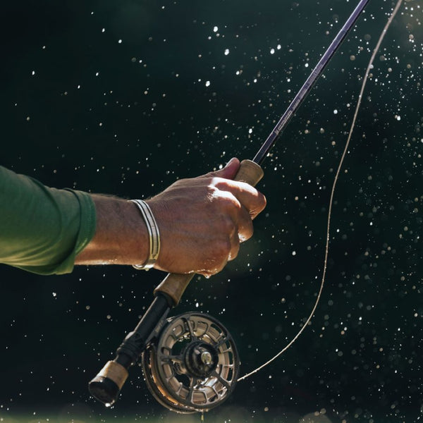 Hand in motion holding a fly fishing rod with the Sage Shift LT Fly Reel and fly line, with water droplets capturing the light against a dark natural background.
