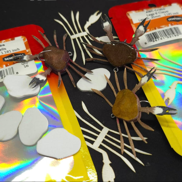 Three crab flies tied with the Semperfli ABC Complete Crab Kits for fly tying, with the packaging on a black background.