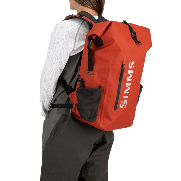 Person in waders wearing the Orange 30-Liter Simms Dry Creek Rolltop Backpack with black straps and Simms Logo on a white background.