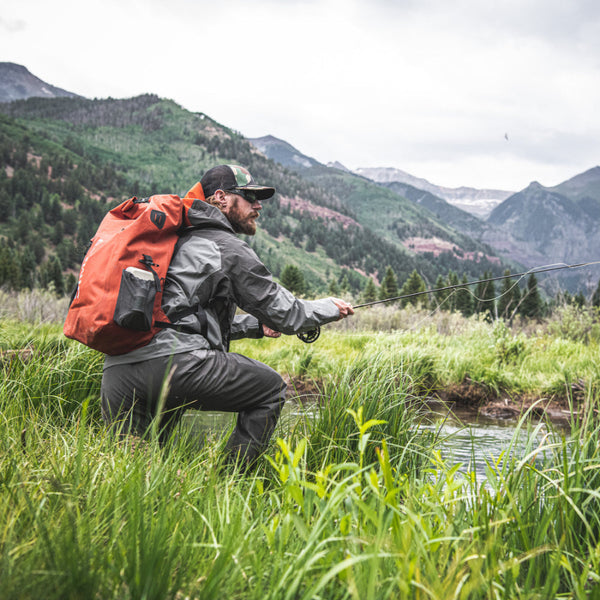 Man with an orange Simms Dry Creek Rolltop Backpack fishing in a mountainous area.