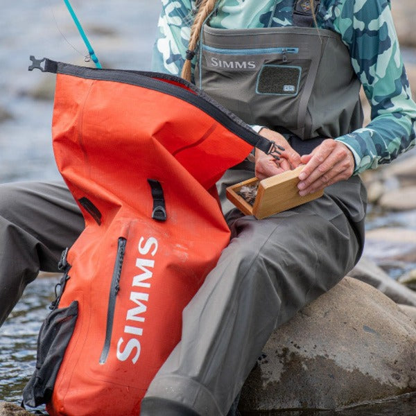 Person wearing waders sitting by a river with an orange Simms Rolltop Dry Creek Backpack.