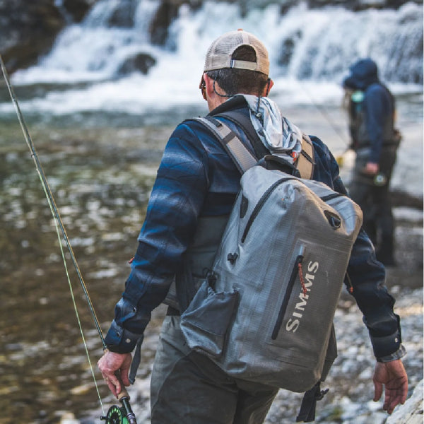 Angler with waders and a fly fishing rod, and wearing the Simms Dry Creek Z Waterproof Backpack, with another angler and a waterfall in the background.