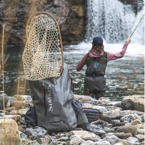 Angler in waders fly fishing in front of a waterfall, with a net and the Simms Dry Creek Z Waterproof Backpack on a gravel bar in the foreground.