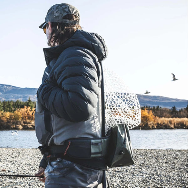 Person with fishing gear wearing waders and the Simms Dry Creek Z Waterproof Waist Pack, surveying the river and opposing bank from a gravel bar.