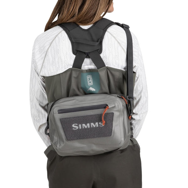 Person wearing waders and the steel grey Simms Dry Creek Z Waterproof Waist Pack, showing the Simms logo on the front pocket, waterproof zippers, and optional neck strap, against a white background.