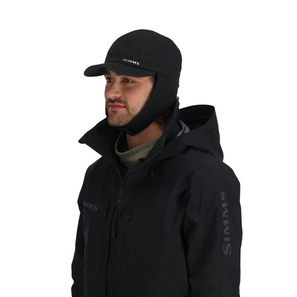 Man wearing a black hooded jacket and black waterproof Simms ExStream Cap with ear flaps on a white background.