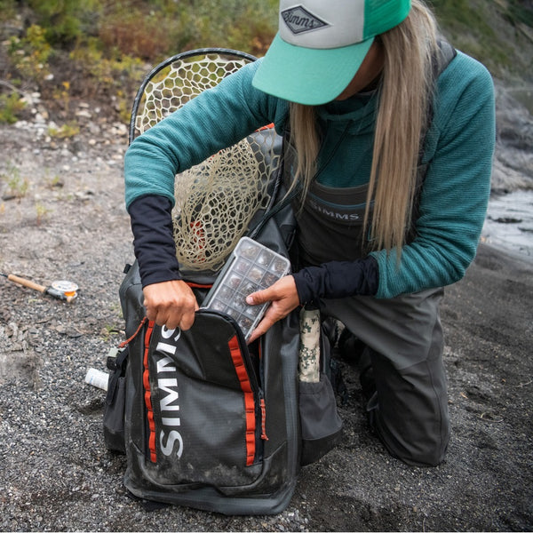 Person on a riverbank loading a grey Simms G3 Guide Rolltop Backpack with fly box, net, and other fishing gear.