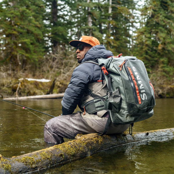 Person with a Simms G3 Guide Backpack fishing on a log over water with trees in the background.