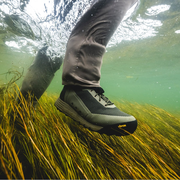 Person wearing wading pants and the Simms Men's Access Wading Boot for fly fishing in dark stone, on a bed of underwater grass.
