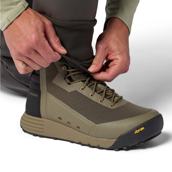 Side view of a person purring on a pair of the Simms Men's Access Wading Boot for fly fishing in dark stone, on a white background.