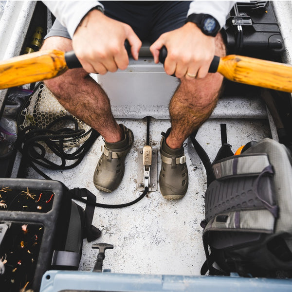 Overhead view of a person rowing a drift boat with square-top wooden oars and wearing black shorts and dark stone brown and beige Simms Men's Drifter AT Fishing Shoe.