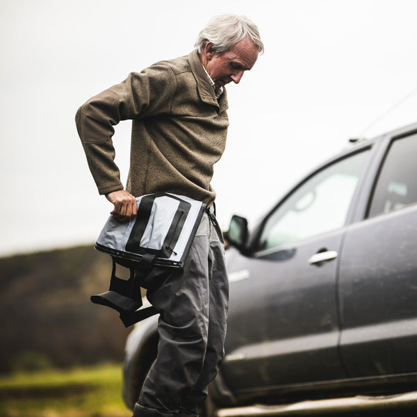 Man standing next to a pickup truck putting on Simms G4Z Fly Fishing Waders over a Simms Harbor Sweater in sturgeon heather.