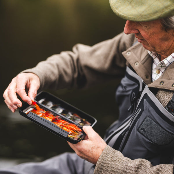 Man sitting next to a river in Scotland wearing Simms G4Z Fly Fishing Waders over a Simms Harbor Fleece Sweater, choosing a bright orange fly from an open fly box in his hands.