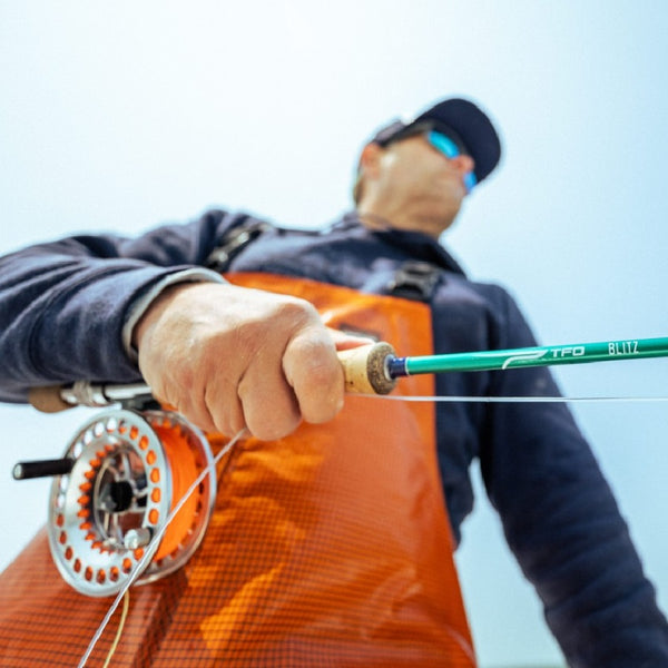 Person in orange fishing bibs holding a TFO Blitz Fly Fishing Rod with reel and line against a cloudy sky.