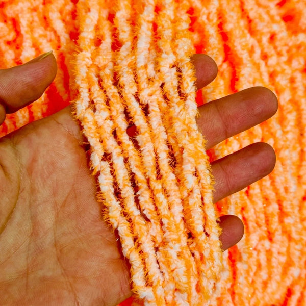 A close-up of a hand holding several skeins of fluffy Upavon Atomic Egg Chenille in hazard orange, with more of the same material visible in the background.