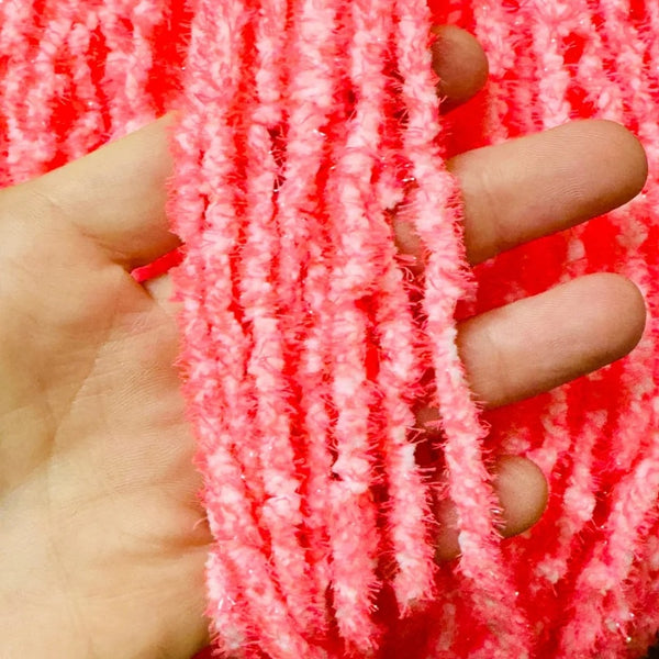 A close-up of a hand holding several skeins of fluffy Upavon Atomic Egg Chenille in isotope pink, with more of the same material visible in the background.