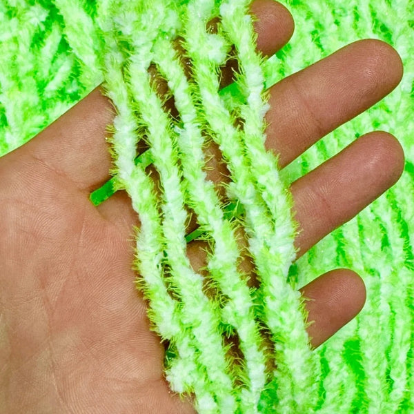 A close-up of a hand holding several skeins of fluffy Upavon Atomic Egg Chenille in toxic green, with more of the same material visible in the background.