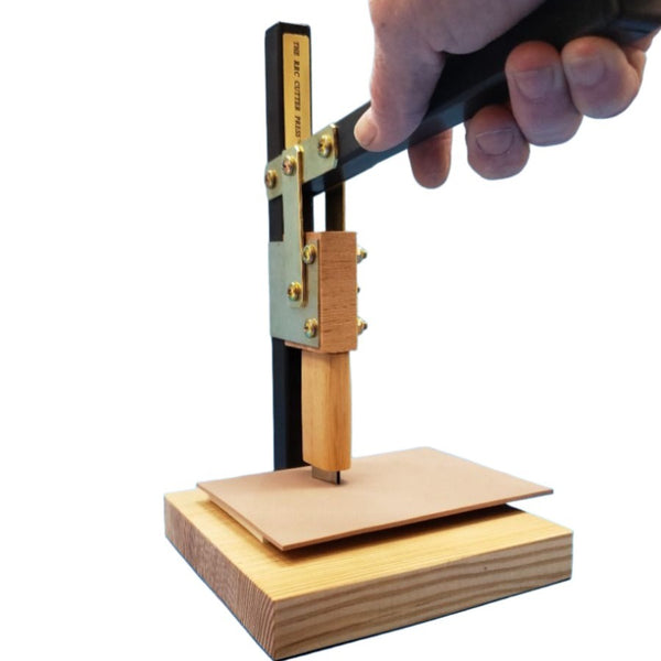 Hand using the lever to begin the cutting action of the River Road Creations Cutter Press Assistive Device, showing a cutter pressing into fly tying sheet foam to cut a specific shape, against a white background.