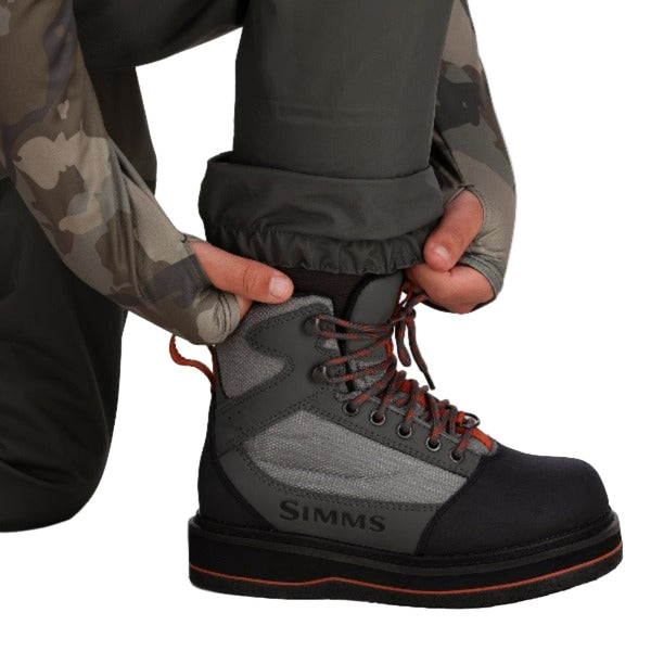 A person kneeling forward wearing Simms Kids' Tributary Stockingfoot Waders zoomed in on the leg and gravel guard details on a white background.