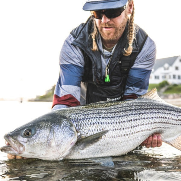 Man wearing the Simms Fall Run Vest over a plaid shirt and holding a large striped bass by a body of water with houses in the background.